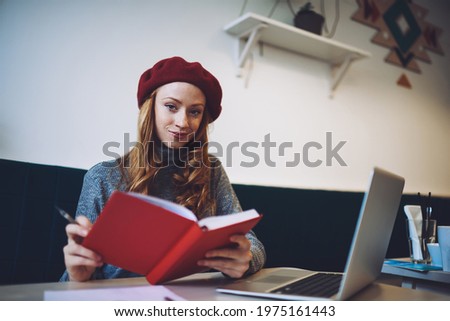 From below of glad female in casual clothes with beret looking at camera sitting at table with laptop and notebook during work