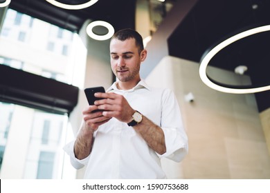 From Below Glad Man In Trendy White Shirt Browsing Smartphone While Standing In Hall Of Urban Business Center On Blurred Background