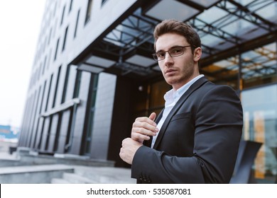 From Below Image Of Business Man In Suit And Glasses Standing Sideways And Looking At Camera Near The Office