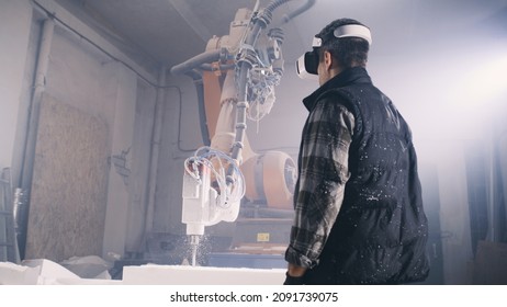 From Below Male Worker Putting On Futuristic VR Goggles And Examining Virtual Plans While Standing Near Robotic Arm Milling Machine In Illuminated Workshop