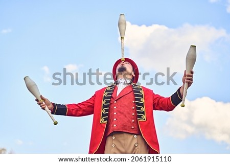 From below man in red costume balancing club on nose while juggling against cloudy blue sky during performance in park