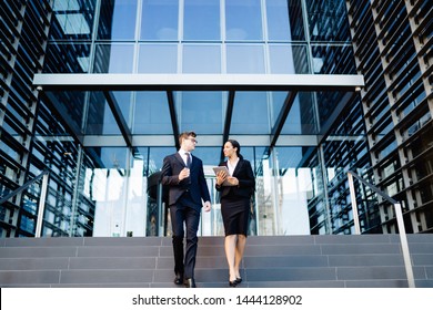 From Below Of Multiethnic Woman And Man In Suits Holding Coffee Cup And Tablet Walking Down Steps Of Office Building And Talking