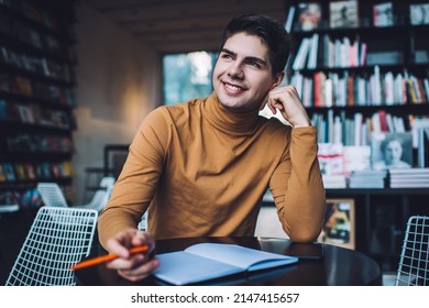 From below of optimistic young male in casual wear sitting at table with notebook in library and smiling while looking away - Shutterstock ID 2147415657