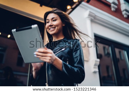 From below pleased Asian woman wearing leather jacket using modern tablet with interest while standing on city street and smiling