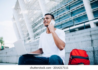 From below of positive African American male freelancer in casual clothes communicating on smartphone while sitting with laptop on knees on stairs near modern building in daytime