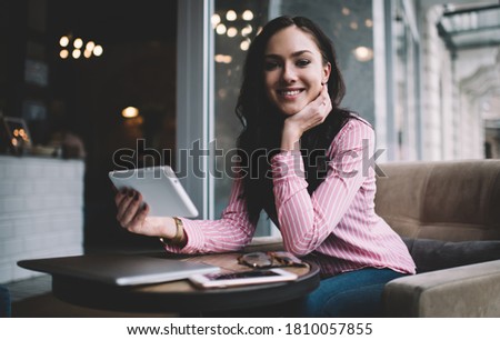 From below of positive female with tablet and laptop with mobile phone sitting in street cafe and looking at camera