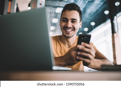 From below of positive man in casual clothes using cellphone chatting with friend browsing laptop sitting at table at workplace