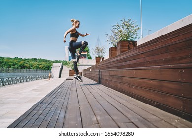 From below side view of active slim motivated female athlete jumping up with high knee lifting on wooden steps of city promenade in summer day
