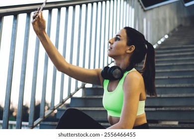 From below side view of athletic female in trendy sports bra and leggings sitting on metal staircase and taking selfie on smartphone while relaxing after training on street - Powered by Shutterstock