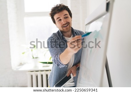 From below of smiling male standing with tablet and marker in hands while pointing at writings on white board and explaining project requirements to unseen audience in room Foto stock © 