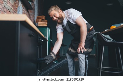 From Below Of Smiling Plus Size Man In Apron Having Fun While Putting Baking Pan Into Oven And Cooking In Kitchen At Home