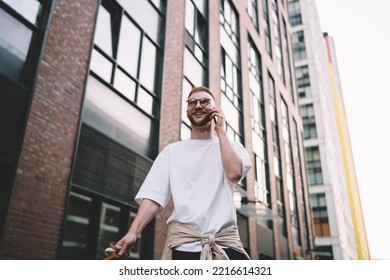 From Below Of Smiling Young Bearded Man With Red Hair In Casual Clothes Making Phone Call While Walking Dog On Leash Near High Rise Urban Buildings