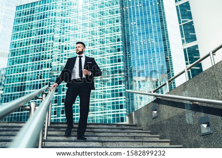 From below of successful businessman in suit walking down stairs in hurry looking away in front of giant modern business center 