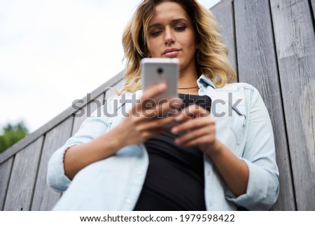 Below view of attractive Caucaisan blogger 20s using modern cellular device for online messaging, hipster girl reading text publication while browsing media content in mobile social networks