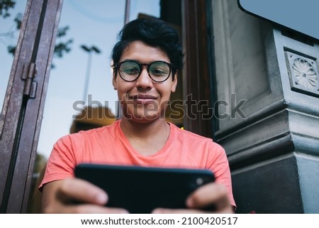 Below view of carefree Latin blogger using 4g wireless internet connection on blurred palmtop technology, handsome guy in optical eyeglasses reading web publication on modern mobile device