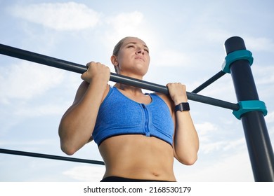 Below view of concentrated powerful young woman with fitness tracker hanging on horizontal bar while doing pull-up at workout area - Powered by Shutterstock