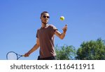 From below view of man in sunglasses practicing forehand shot hitting yellow ball with racket on tennis court