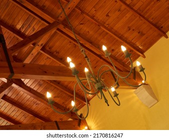 Below view of an old fashioned chandelier with candle lightbulbs hanging from sturdy wooden roof ceiling inside. Architecture beam detail, interior design, brass, antique light fixture in empty room