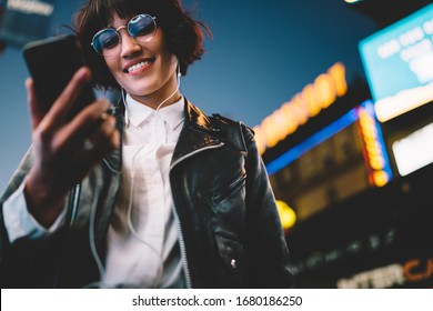 Below view of pretty woman in cool eyeglasses and trendy wear smiling on metropolitan street with night lights enjoying songs from playlist in earphones, girl reading sms with good news on smartphone - Powered by Shutterstock