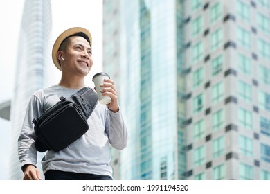 Below View Of Smiling Young Vietnamese Man With Belt Bag Listening To Music In Wireless Ear Buds And Holding Coffee Cup While Walking Along Street With Skyscrapers