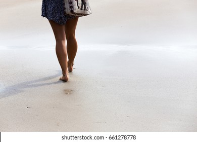 Below Waist Shot Of A Toned Girl's Legs Walking On A Bright Sunny Beach With Barefoot As A Bag Hangs From Her Shoulder.