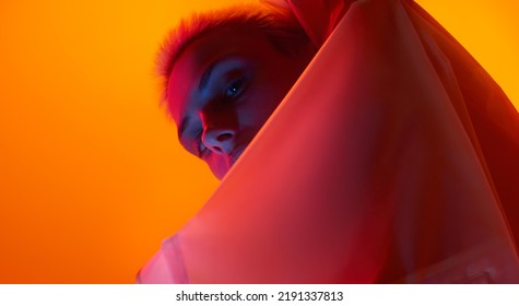 From below young female model looking at camera from behind futuristic apparel while standing under red neon light against orange background - Powered by Shutterstock