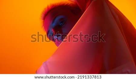 From below young female model looking at camera from behind futuristic apparel while standing under red neon light against orange background
