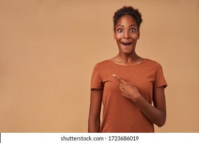 Bemused young pretty dark skinned curly woman looking surprisedly at camera with wide eyes and mouth opened while pointing aside with forefinger, isolated over beige background - Shutterstock ID 1723686019