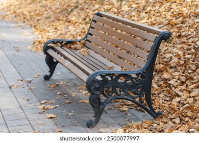 A bench with a black and brown frame sits on a brick walkway. The bench is empty and surrounded by fallen leaves. Concept of tranquility and peacefulness, as the bench is a quiet spot to sit