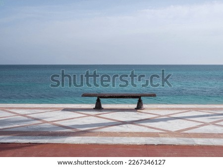Bench made of wood, standing on a pathway covered in orange tiles. The bench is overlooking a beautiful seaside landscape with no people around. 