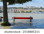 A bench is in the water. River Danube flooding in Budapest, 2024 autumn. The lower quay is under water. Buda side with old buildings in the background