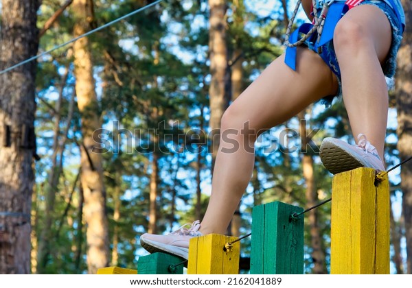 Bending her knees, the girl in pink sneakers slowly\
and carefully walks along the bars of the rope path. A woman in\
short shorts and with beautiful legs passes an obstacle course in\
an extreme park