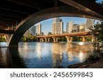 Beneath the North Lamar Bridge in Austin, Texas, the tranquil waters of Lady Bird Lake mirror the golden hues of the cityscape, creating a beautiful reflection on a summer day.