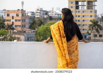 A Bengali woman standing on roof of house in yellow saree looking away. Saree is traditional and popular garment for women in Bangladesh. Backside view. - Powered by Shutterstock