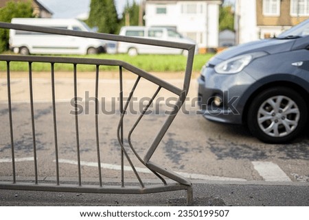 Bent railing metal fence smashed in by car grey small vehicle on cement pavement road street carriageway near crosswalk walkway with grass houses and vans in background painted lines sunny shade crash