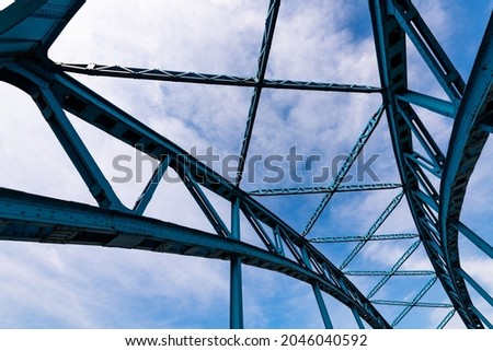 Bent steel river bridge with bright blue cyan color in Duisburg Ruhr Basin Germany. Construction with rivets, fixings, beams from wide angle frog perspective on a sunny blue sky day.