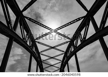 Bent steel river bridge in Duisburg Ruhr Basin Germany. Construction with rivets, fixings, beams from wide angle frog perspective on a sunny day. Black and white greyscale with sun in the middle.