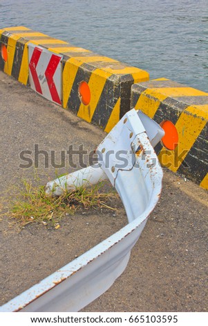 A bent and twisted white metal guardrail and yellow and black concrete blocks with orange safety reflectors mark an extremely dangerous curve on a road beside the Pacific Ocean in Japan.