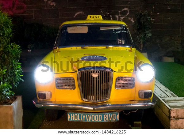 BERGEN, NORWAY - MAY,\
2019: Old american yellow taxi with headlights on at night as a\
decoration next to a cafe \