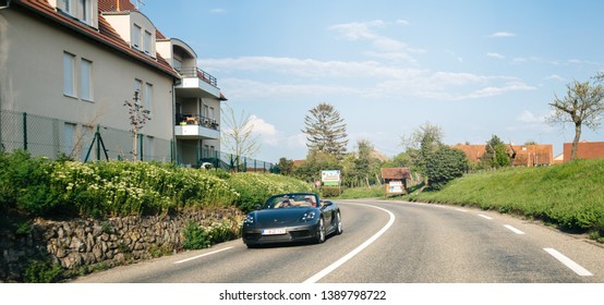 Bergheim, France - Apr 19, 2019: Couple Driving Fast In Their Black Porsche Convertible Car On The Village Road Of Bergheim