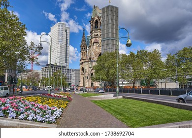 Berlin Germany, 10.05.2020: The Kaiser Wilhelm Memorial Church, In German Kaiser-Wilhelm-Gedächtniskirche, Spring Time In The City, With A Blue Sky And Colourful Flowers, Green Trees On The Road Side