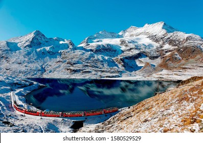 A Bernina Express train traveling along the lake shore of Lago Bianco and Piz Cambrena towering under blue sky in background after a snowfall in autumn, near Ospizio Bernina, in Grisons, Switzerland - Shutterstock ID 1580529529