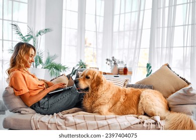 Best animal companion. Side view of charming young woman lying on comfy couch with adult golden retriever and working on wireless laptop. Freelancing at home with favorite pet.