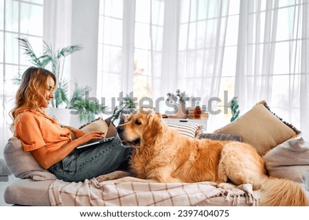 Best animal companion. Side view of charming young woman lying on comfy couch with adult golden retriever and working on wireless laptop. Freelancing at home with favorite pet.