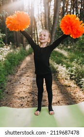 The Best, Beautiful, Smiling Girl, A Professional Athlete, A Child Cheerleader In A Black Suit Stands On A Pedestal Raises His Hands Up With Orange Pom-poms, Victory. Cheerleading Champion.