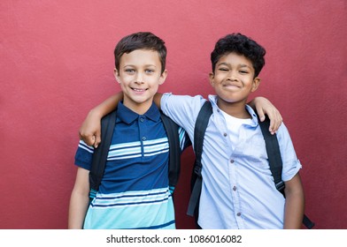 Best Children Friends Standing With Hand On Shoulder Against Red Background. Happy Smiling Classmates Standing Together On Red Wall. Portrait Of Multiethnic Schoolboys Enjoying Friendship.