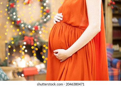 Best Christmas Gift. Faceless Pregnant Woman In A Red Dress Hugs Her Belly Against The Background Of The Christmas Living Room In Blur. Close-up No Face.