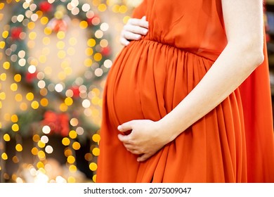 Best Christmas Gift. Pregnant Woman In A Red Dress Hugs Her Belly Against The Background Of The Christmas Living Room In Blur. Close-up No Face.