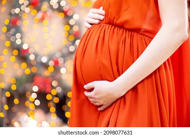 Best Christmas Gift. Pregnant Woman In A Red Dress Hugs Her Belly Against The Background Of The Christmas Living Room In Blur. Close-up No Face.