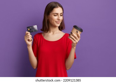 Best coffee in app. Happy young pretty brunette girl in red t-shirt holds disposable cup and showing a debit or credit card. Pays by internet for hot drink. Posing over purple wall - Shutterstock ID 2209121385